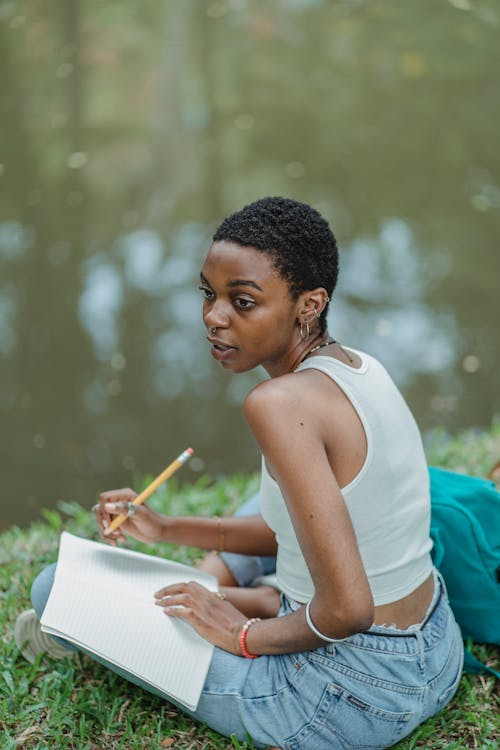 Full length of young African American lady with diary and pencil sitting near lake on green lawn in summer day while looking away