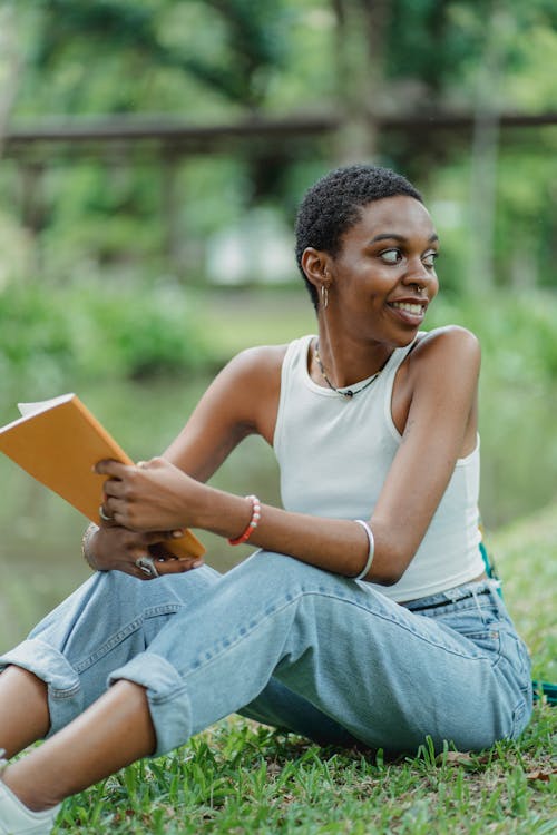 Young African American woman sitting near lake on green grass with sketchpad in summer day and wearing casual outfit while looking away