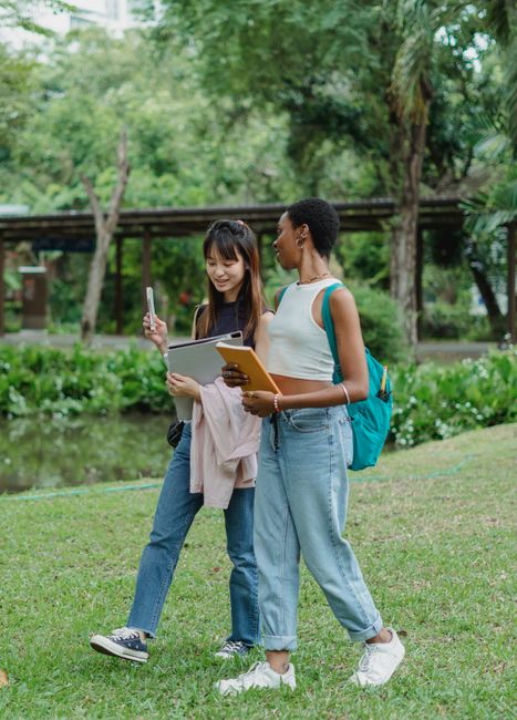 Full body young multiethnic women talking to each other wearing casual summer clothes with backpack while walking on grass notebooks