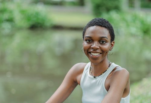 Free Smiling young African American female sitting near lake on green grass in summer day while looking at camera Stock Photo