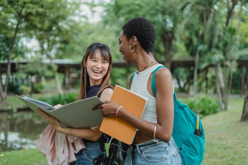 Young multiracial women in casual summer outfits with backpack speaking with each other while walking on grass with notepad and folder and looking at each other