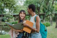 几位穿着休闲夏装、背着背包的年轻多族裔女性，一边走在草地上，一边拿着记事本和文件夹交谈，同时互相看着对方