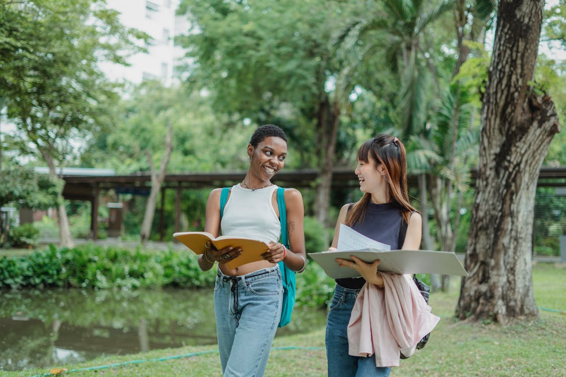 Smiling young multiethnic women talking to each other while walking in summer park sharing notes and looking at each other