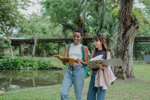 Smiling young multiethnic girlfriends speaking with each other while walking in green park with backpack and notepads in summer day