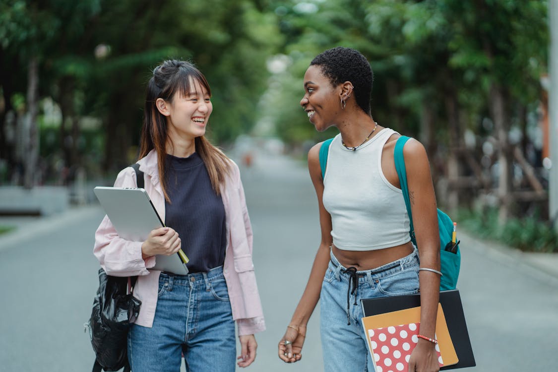 Smiling young multiethnic women standing in middle of street with laptop and notebooks while speaking with each other in summer day and wearing casual clothes while looking at each other