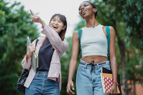 Cheerful Asian schoolgirl with laptop and copybooks pointing away while strolling together with smiling African American friend in lush summer park