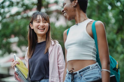 Optimistic smiling diverse female students wearing casual outfits with backpack and laptop strolling together in lush park and discussing term results while looking at each other happily