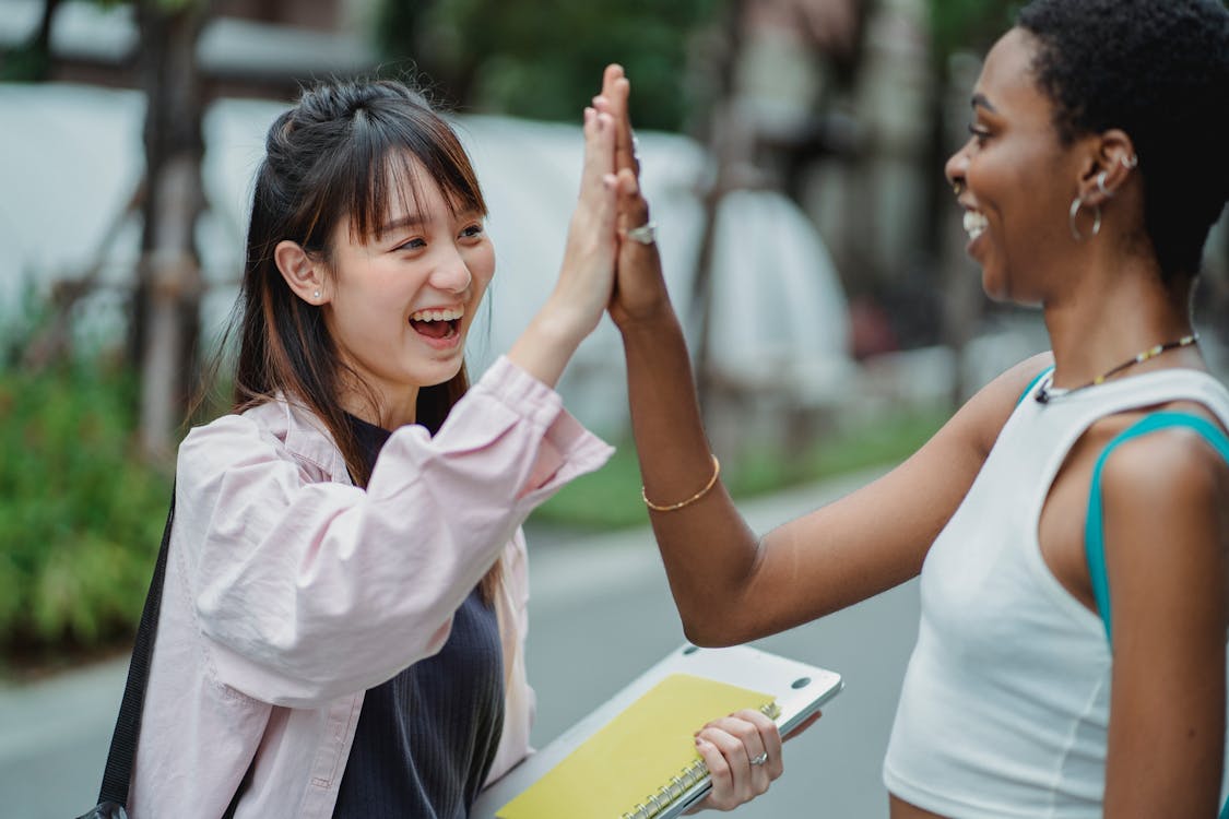 Free Side view excited multiracial female students in casual clothes giving high five to each other while spending break in lush park Stock Photo