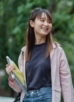 一位年轻的亚洲女子面带微笑，拿着笔记本电脑和书籍，在户外享受阳光明媚的一天