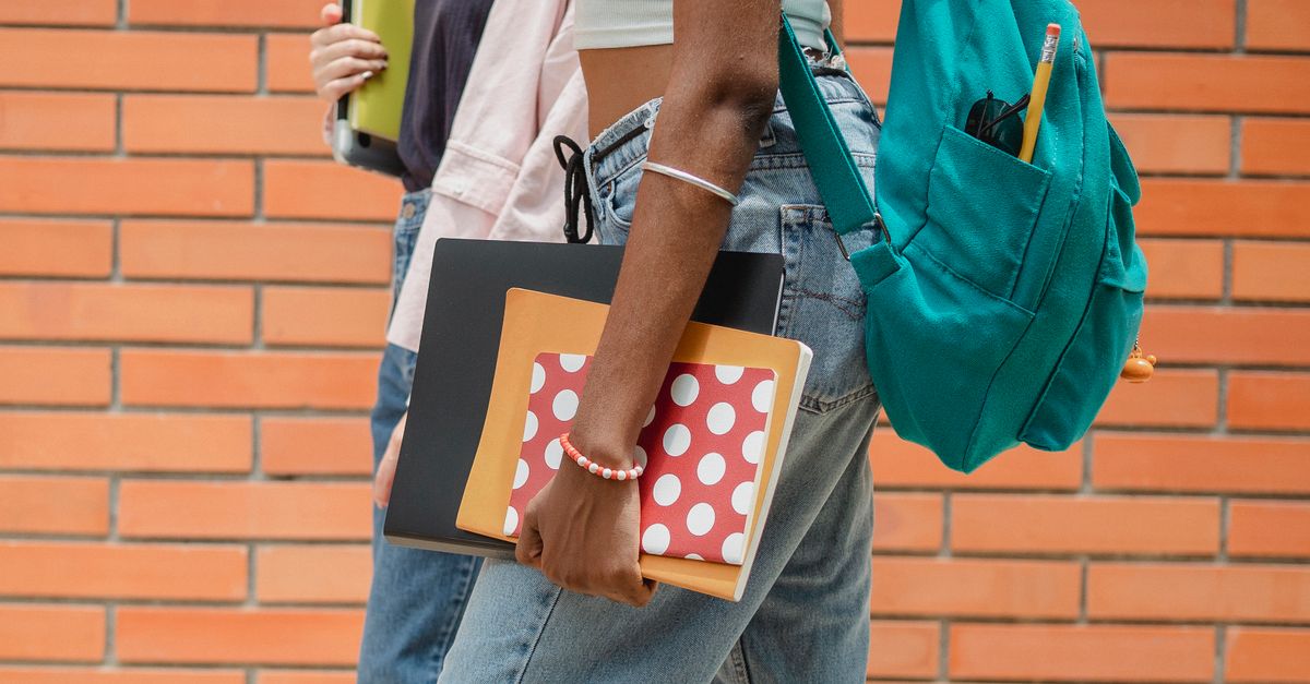 Side view crop female students in casual clothes with backpacks and copybooks strolling together against brick wall
