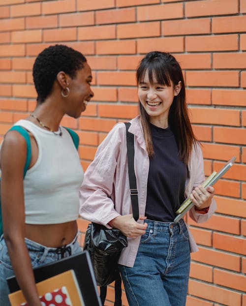 Free Laughing multiethnic female students wearing casual outfits strolling together against brick wall and chatting happily while looking at each other with smiles Stock Photo