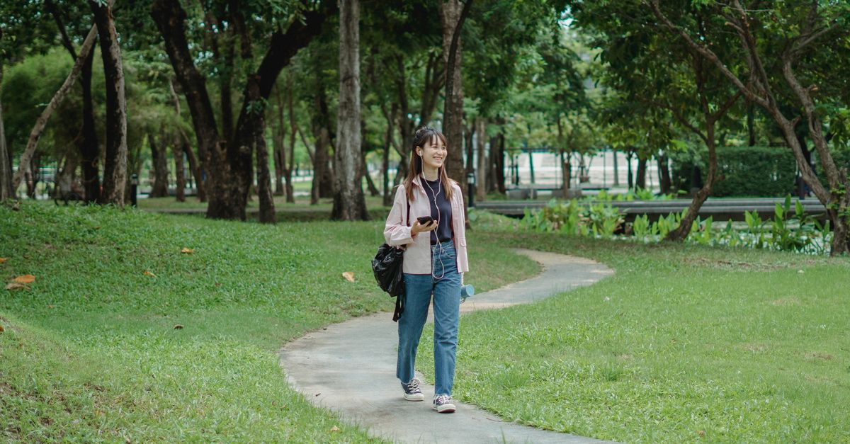 Full body of positive young Asian female in casual outfit listening to music via earphones and smartphone while walking on pathway in park