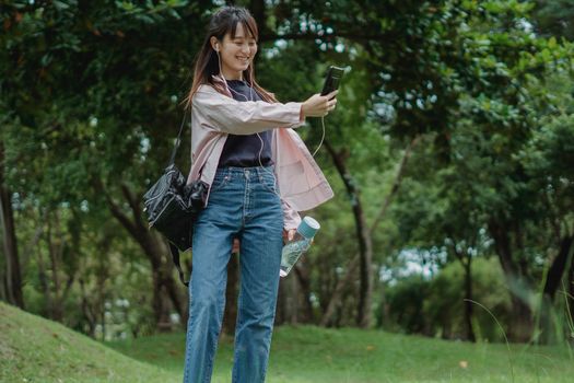 一位穿着休闲服装的快乐年轻亚洲女性在绿色公园里边散步边用耳机打视频电话