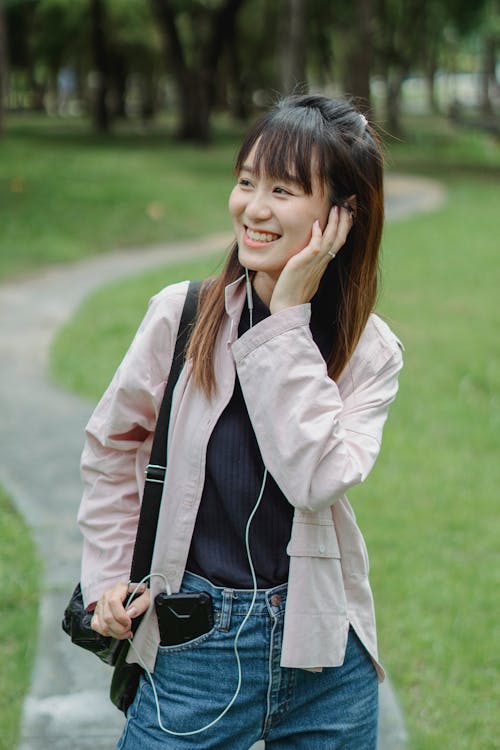 Cheerful young Asian female in casual clothes standing on pathway in park while walking and listening to music via earbuds