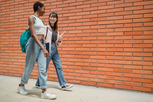 Full length of cheerful multiracial young female students strolling together near brick wall and looking at each other while having conversation