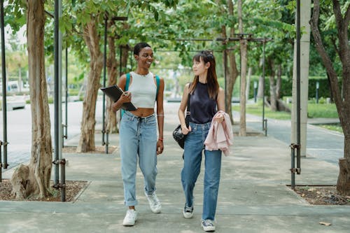 Young multiethnic female friends strolling along walkway between trees and talking to each other