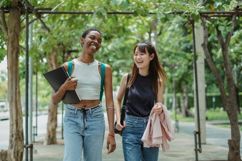 Free Positive young multiracial female friends in casual outfits with backpacks having conversation while walking along alley Stock Photo