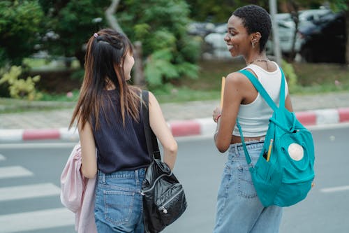 Free Back view of young multiracial female friends in casual outfits with backpacks laughing on crosswalk Stock Photo
