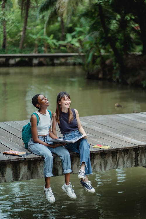 Cheerful multiethnic female friends in casual wear sitting on wooden bridge with copybook and laughing