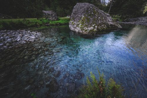 Free Huge triangular boulder covered with moss in narrow blue river surrounded by verdant trees in forest in daytime Stock Photo