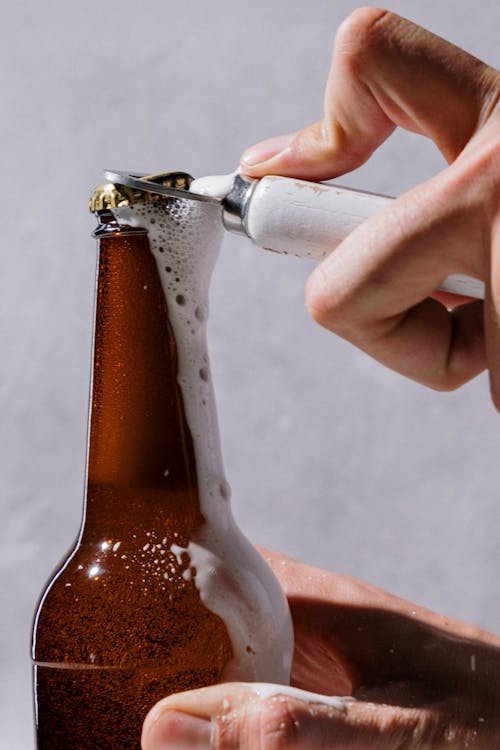 Foto de uma mão abrindo uma garrafa de cerveja. A espuma está saindo, a imagem dá um pouco de sede.