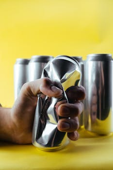 A hand crushing a silver beverage can against a yellow background, symbolizing strength.
