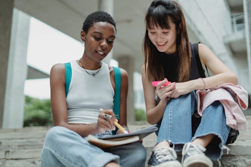 Free Young smiling diverse friends in casual outfits sitting on concrete stairs and taking notes on notepad Stock Photo