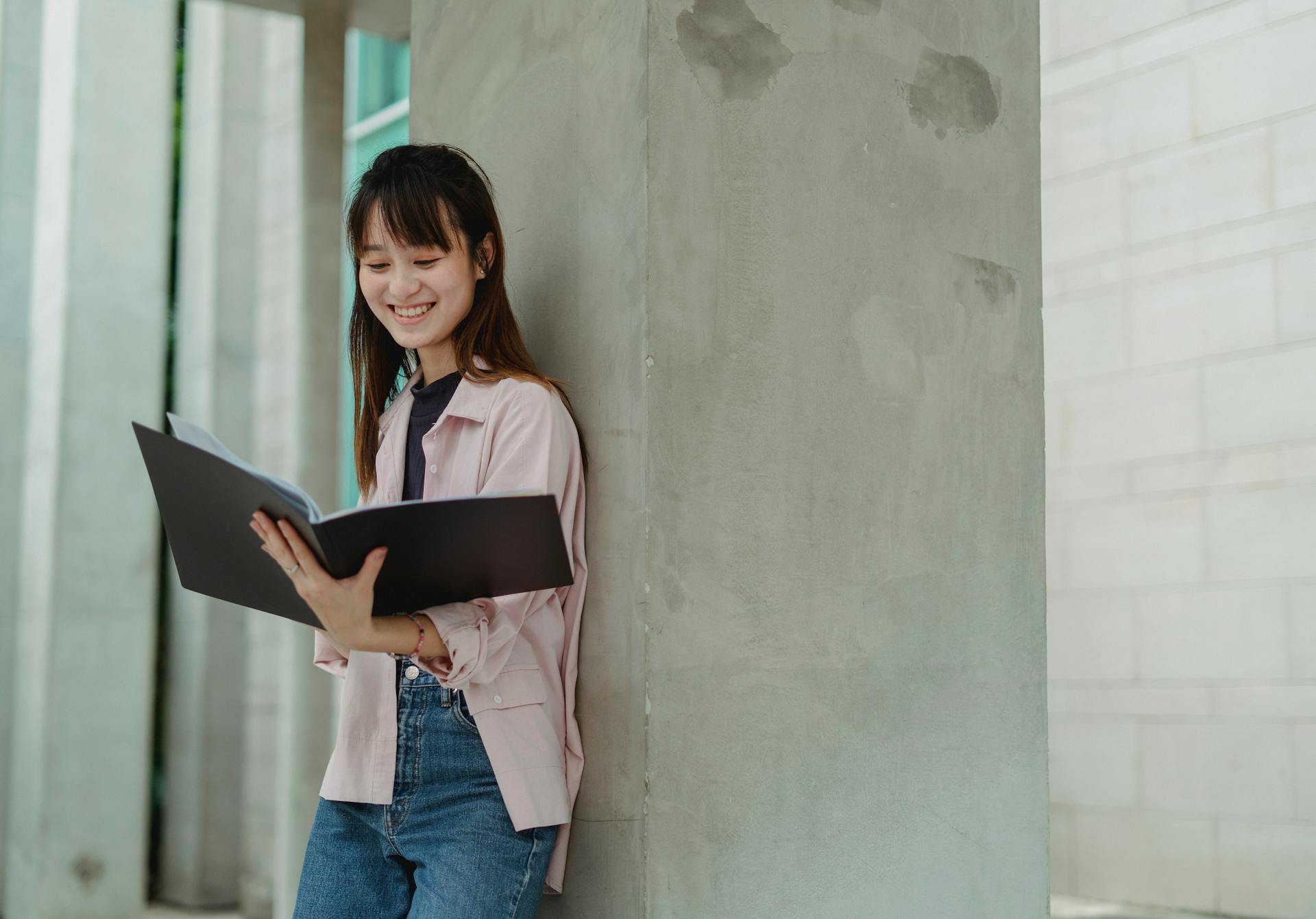 Young Asian female student in casual outfit smiling while reading papers in folder standing near column