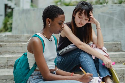 Free Focused young multiethnic female friends sitting on concrete stairs and thinking about project taking notes in notepad Stock Photo