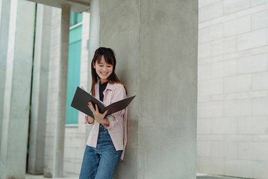 一位穿着休闲衣服、面带微笑的年轻亚洲女学生倚靠在柱子上，阅读文件夹里的文件