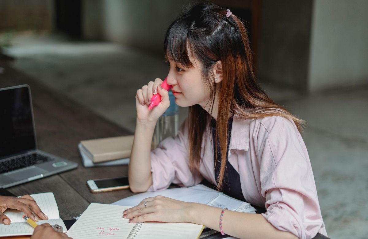 Side view of thoughtful Asian female student sitting at wooden table and taking notes while working on project