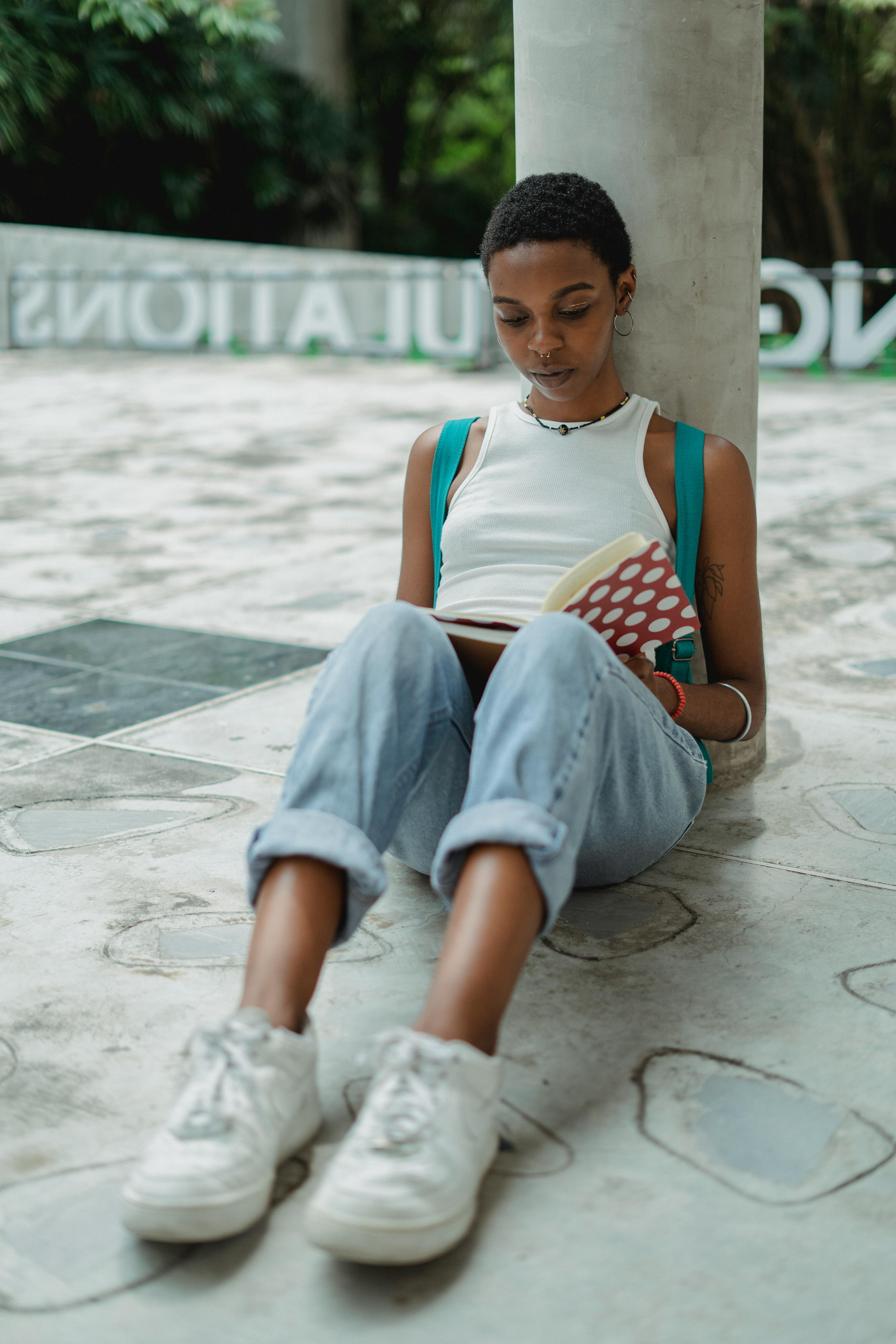 Free Focused young African American female student in casual clothes sitting on ground and reading notes in notebook Stock Photo