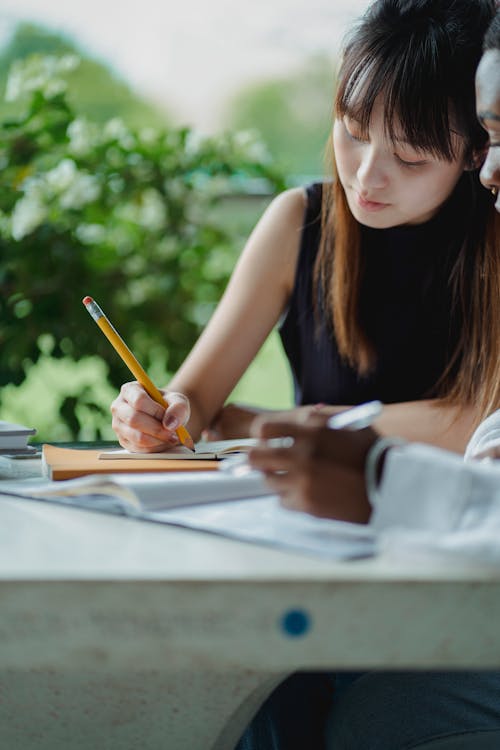 Concentrated Asian female student taking notes while doing research with African American female sittin at table