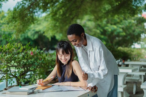 Free Young multiethnic women tutoring with books while spending time outdoors together and wearing casual outfit in summer day at table Stock Photo