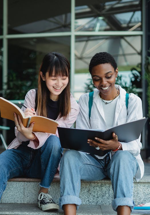Free Smiling young multiracial ladies sitting outside in casual outfit with notebooks and talking with each other in daytime Stock Photo