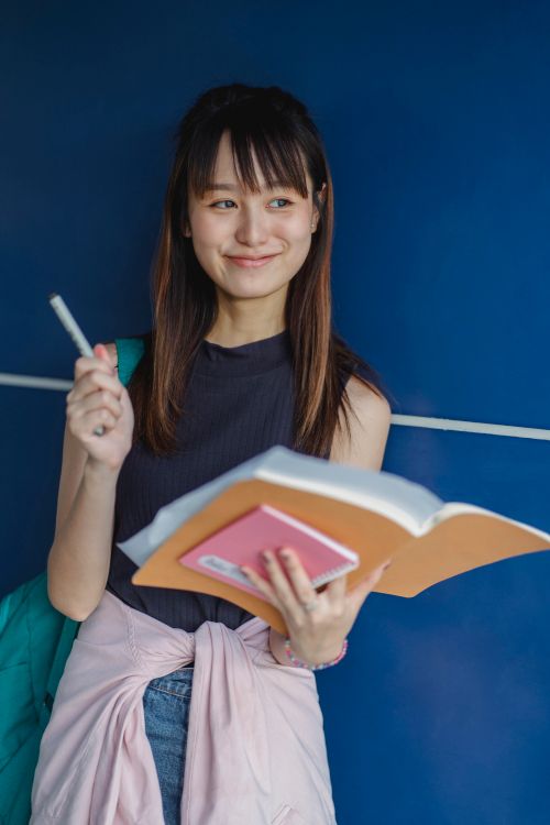 Free Smiling Asian female student in casual outfit standing with notebook and pen on blue background and looking away Stock Photo