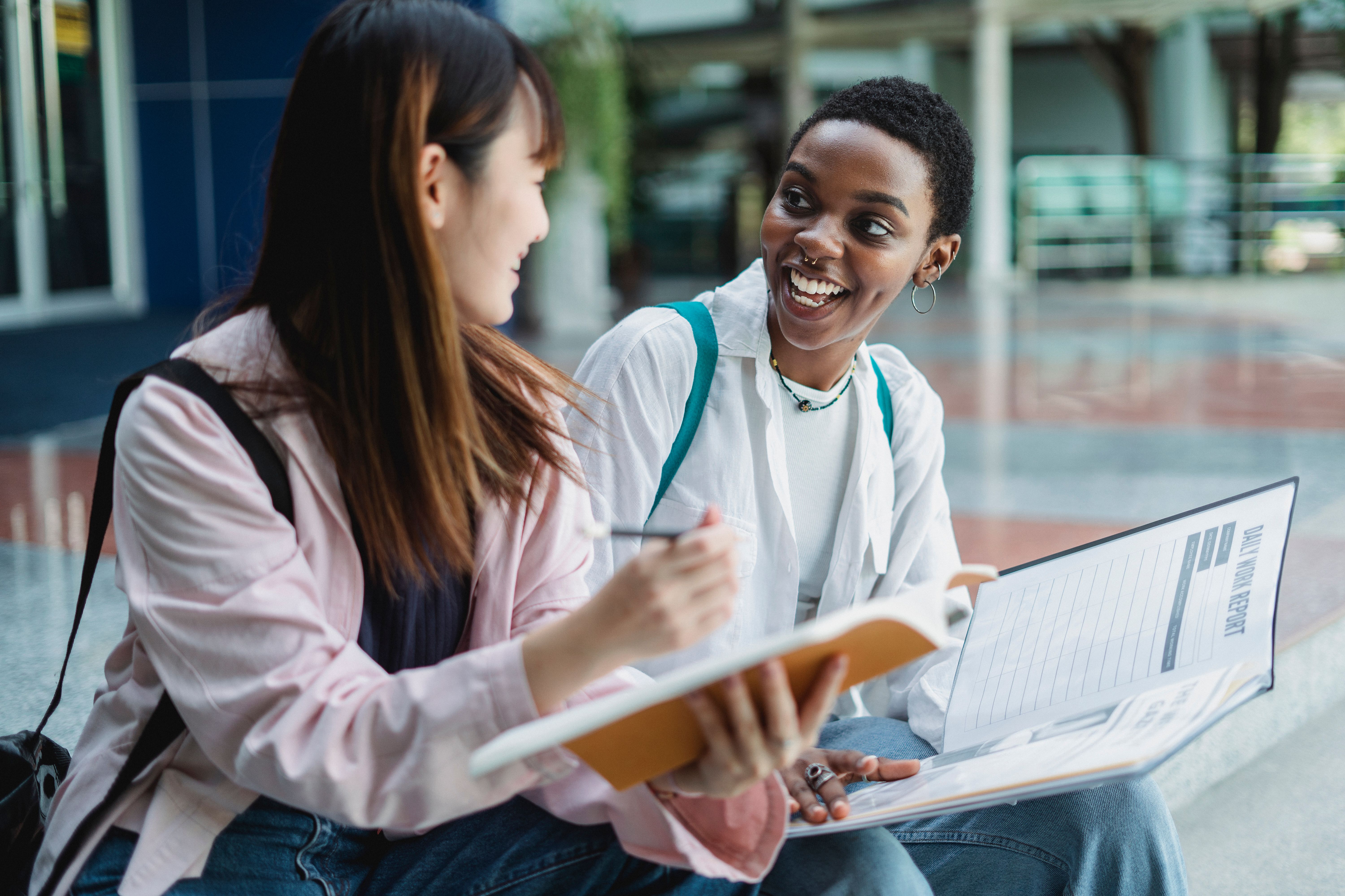 Two students sit together outdoors, smiling while reviewing notes and documents.