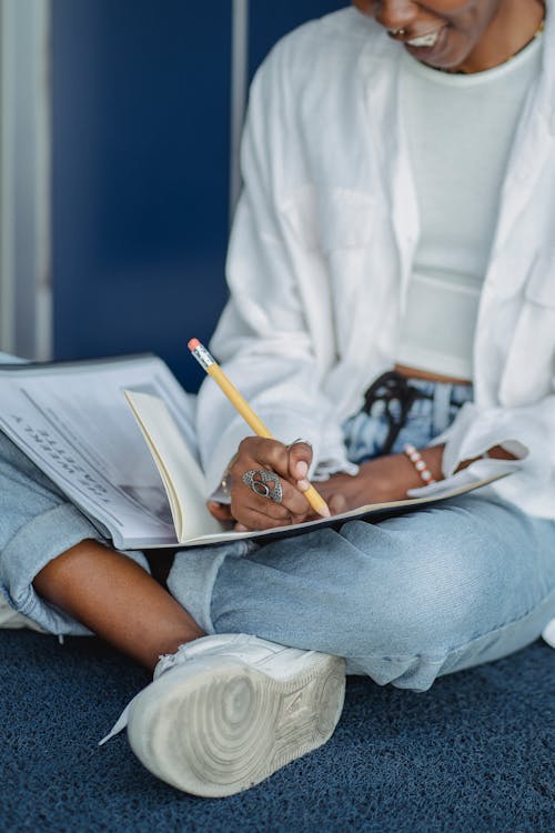 Crop African American female student sitting with crossed legs and writing in notebook while doing homework