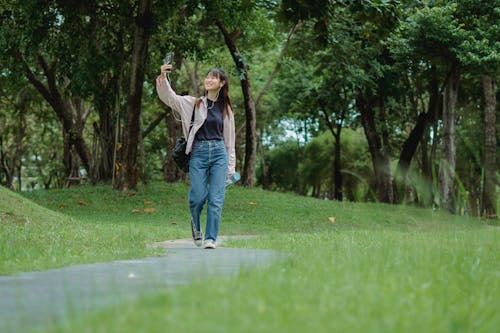 Smiling young Asian woman talking via smartphone while walking in park