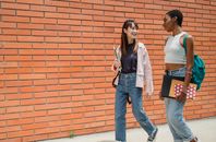 一位背着笔记本和背包的积极非裔美国女学生，在大学砖墙附近散步时，与一位穿着牛仔裤的女性朋友交谈