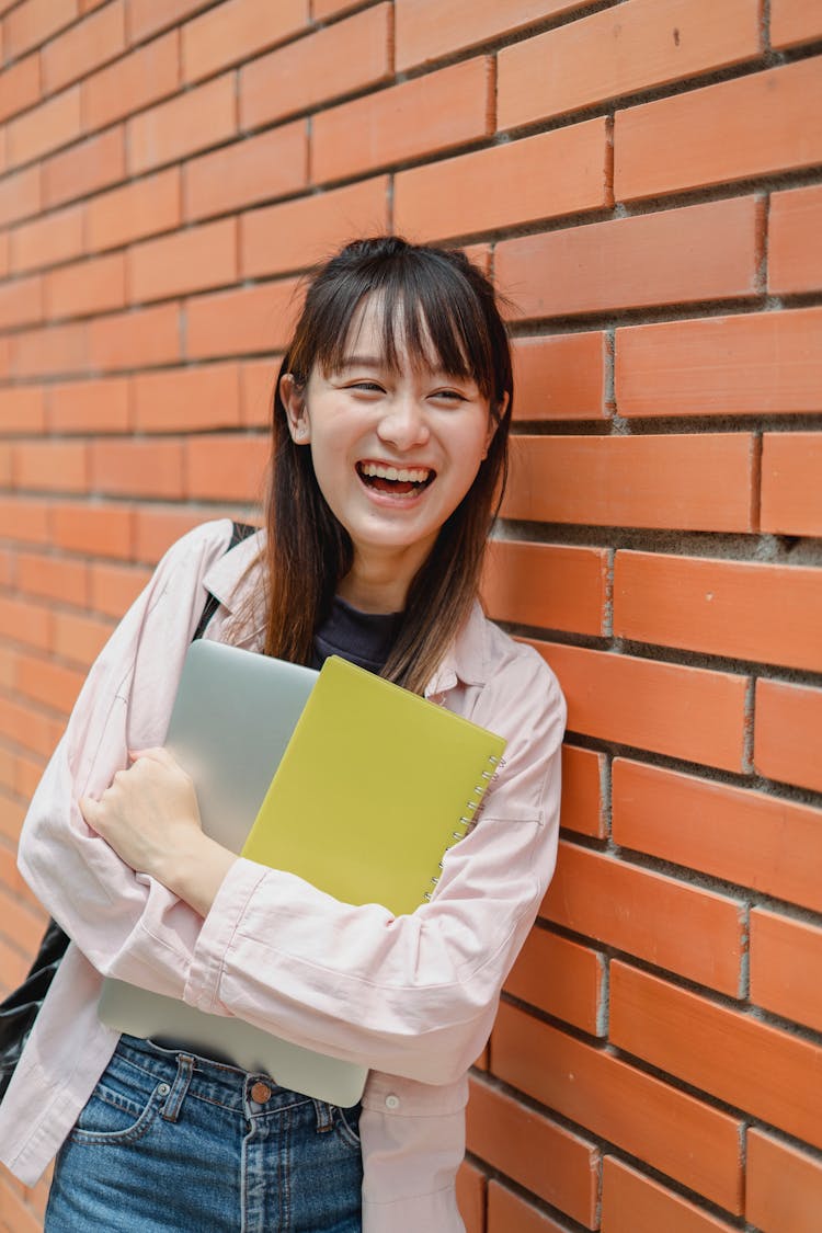 Cheerful Asian Student With Laptop Laughing While Leaning On Brick Wall