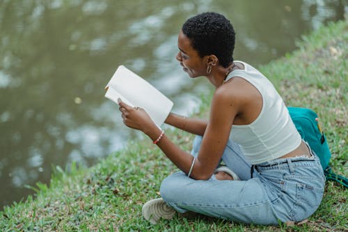 From above of positive young African American female student wearing trendy jeans sitting on grassy bank of lake and reading textbook