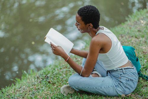 Free From above of positive young African American female student wearing trendy jeans sitting on grassy bank of lake and reading textbook Stock Photo
