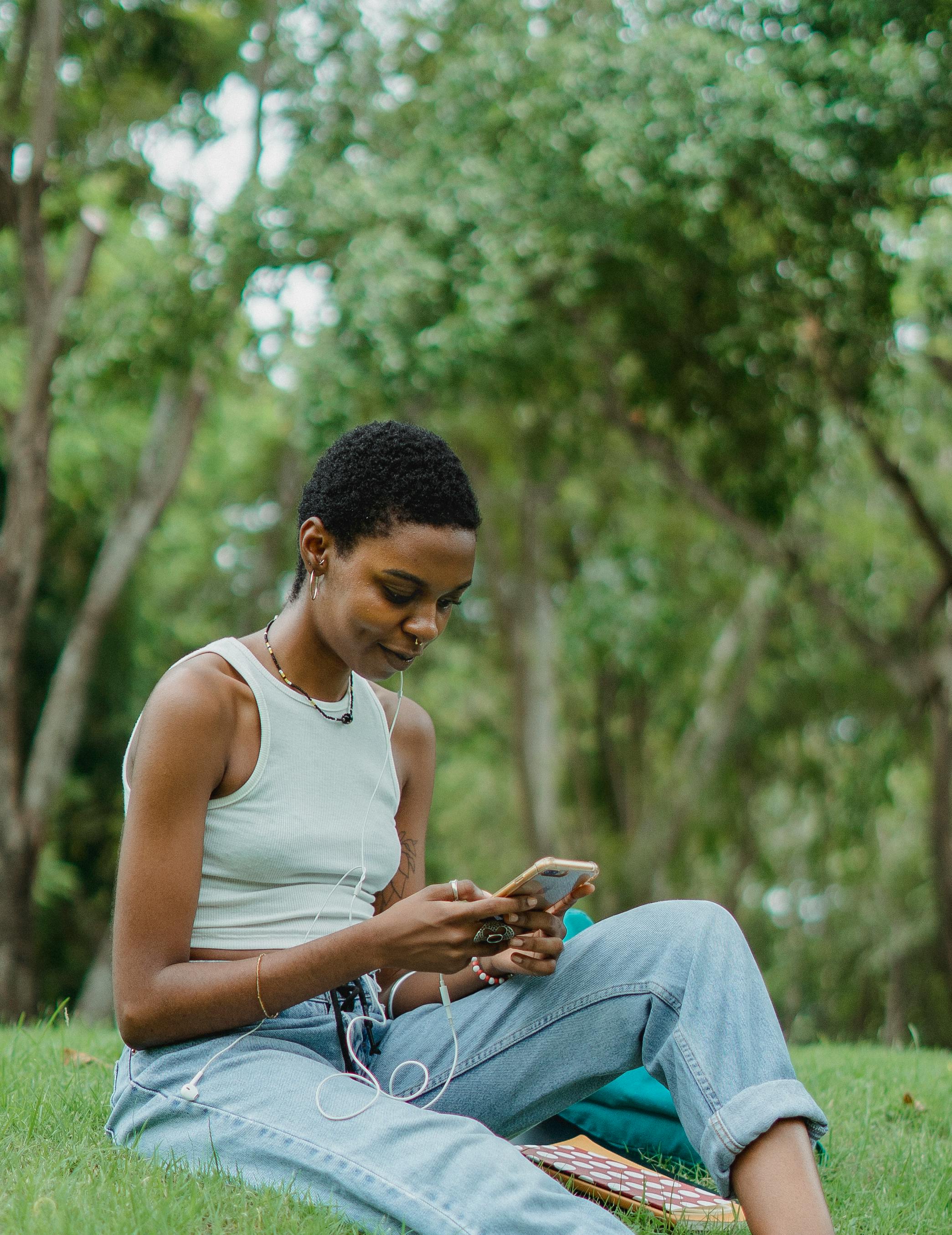 smiling african american young woman sitting on grass and using smartphone