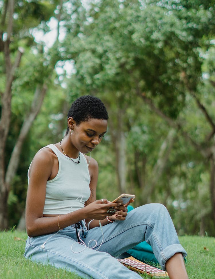 Smiling African American Young Woman Sitting On Grass And Using Smartphone