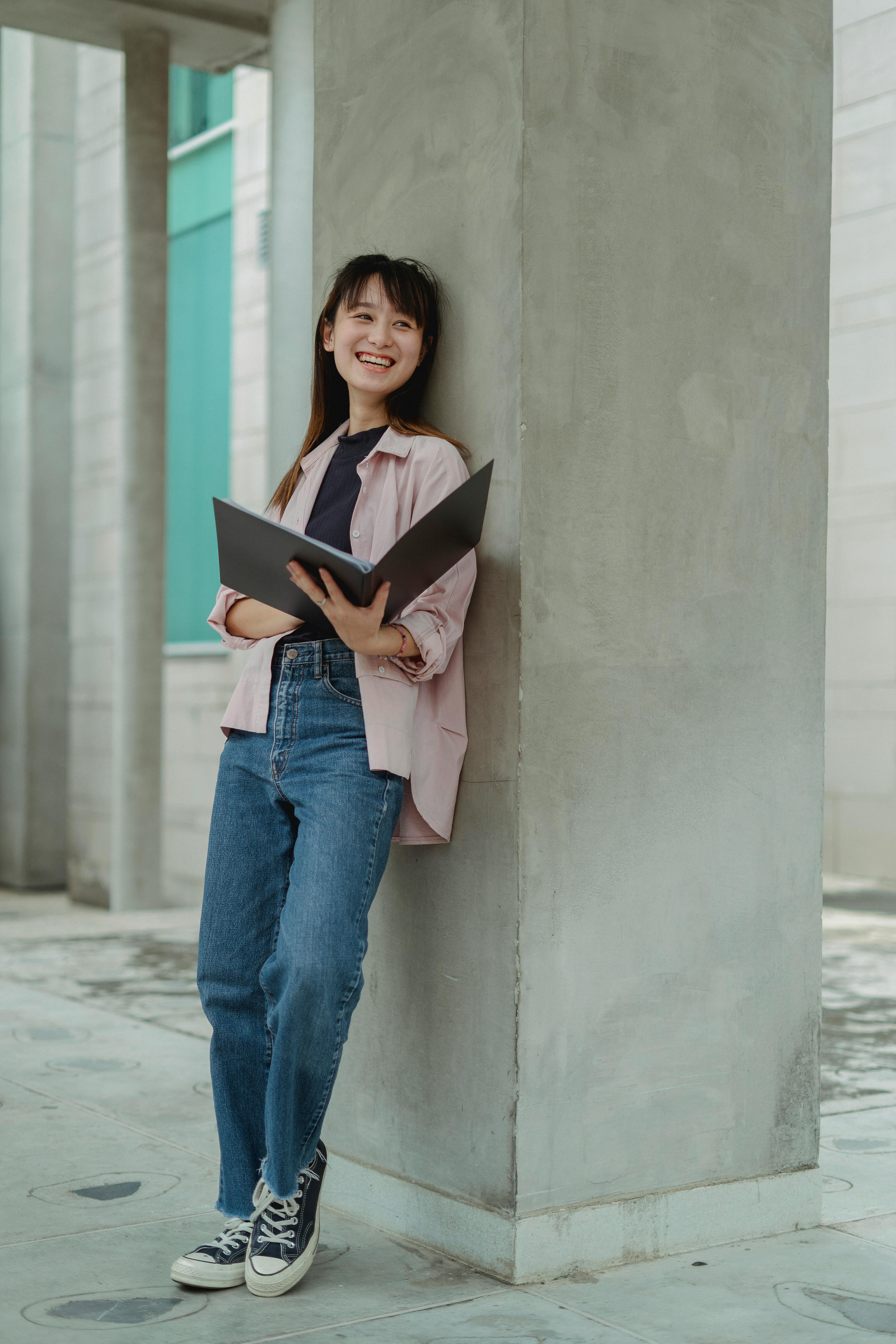 cheerful young woman with folder standing near concrete column