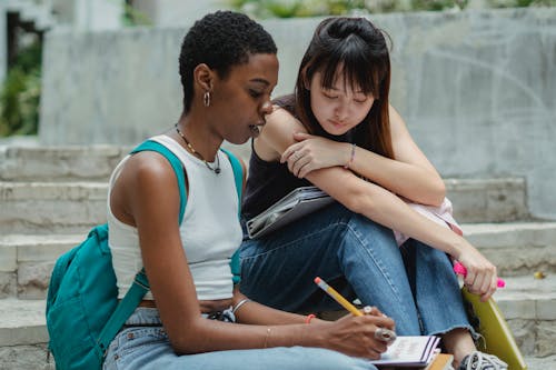 Black female student taking notes while sitting together with Asian friend on concrete stairs and preparing university project