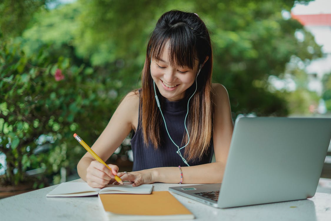 Positive young Asian female student with earphones writing in copybook while doing homework at table with laptop in street cafeteria.