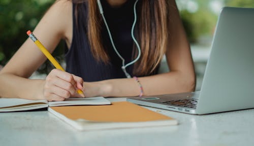 Free Crop anonymous young female freelancer with earbuds writing notes with pencil in notebook while sitting at table with laptop in contemporary cafe Stock Photo