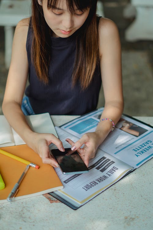 From above of crop focused Asian woman surfing modern smartphone while sitting at table with papers and pens